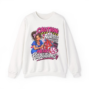 Peut inclure: Un sweat-shirt blanc à manches longues avec une impression graphique représentant une infirmière utilisant un stéthoscope sur un cœur de dessin animé. Le texte indique CAUTION: NURSES MAY CAUSE HEART PALPITATIONS, avec des cœurs roses et un moniteur.