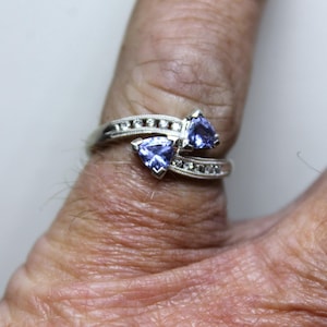 Könnte beinhalten: Ein Ring im Bypass-Stil aus silberfarbenem Metall mit zwei hellvioletten Edelsteinen im Trillantschliff in der Mitte. Die geteilte Ringschiene ist mit kleinen, runden, klaren Steinen besetzt und weist einen feinen Milgrain-Rand auf.