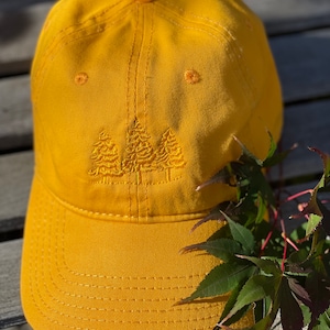 May include: A yellow baseball cap with an embroidered design of three trees. The cap has a curved brim and a stitched design.