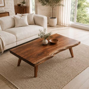 May include: A rectangular coffee table made of dark brown wood with a natural live edge finish. The table features four tapered wooden legs and a smooth, polished surface. A small white ceramic vase and a wooden bowl sit on the tabletop.