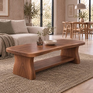 Op de afbeelding: Een lage, rechthoekige salontafel van massief bruin hout met een glad tafelblad met afgeronde randen en een onderste opbergplank. De tafel heeft twee brede, gebogen zijsteunen die het bovenoppervlak met de onderste plank verbinden.