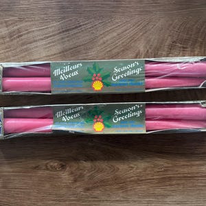 Könnte beinhalten: Zwei Schachteln Bonsoir Stabkerzen in leuchtendem Pink. Jede Schachtel enthält zwei Kerzen und ist mit einem grünen Etikett versehen, das den Text Meilleurs Voeux und Season's Greetings sowie eine Stechpalmenillustration und ein gelbes Muschellogo zeigt.