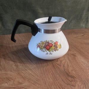 May include: A white teapot with a black handle and lid, featuring a metallic band around the neck. The body displays a colourful botanical illustration of fruits and flowers with the text "Le Thé" printed in black script below the design.