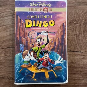 Peut inclure: Une cassette VHS Walt Disney Collection Or intitulée Complètement Dingo, illustrée avec Dingo et Max dans un canyon, une raquette de tennis, un poisson bleu et un radeau jaune, avec le texte Version Française de A Goofy Movie inscrit.