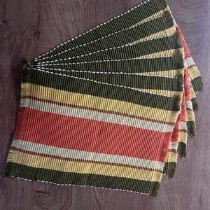 Peut inclure: Un ensemble de sets de table rectangulaires présentant un motif à rayures vert olive, crème et rouge terre cuite. Les sets de table ont une texture côtelée et sont finis avec des bords à franges effilochées, empilés en éventail.
