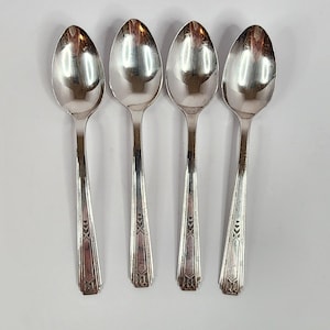May include: A set of four silver-toned metal spoons arranged vertically. Each spoon features a polished oval bowl and a slender handle decorated with a geometric pattern of vertical lines and small triangular accents near the base of the handle.