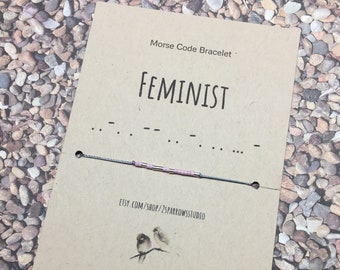 May include: A brown card with the text "Morse Code Bracelet" and the word "FEMINIST" in all caps. Below the word "FEMINIST" is a line of dashes and dots representing the word in Morse code. The card also includes the text "etsy.com/shop/2sparrowsstudio" and a drawing of two birds on a branch.