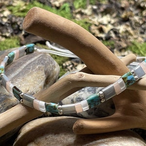 May include: A turquoise, gray, and pink beaded bracelet with silver spacers. The bracelet is made with square-shaped beads and is strung on a white cord.