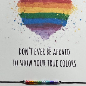 May include: A rainbow heart with a splatter effect and the text "Don't ever be afraid to show your true colors". A rainbow beaded bracelet is below the text.