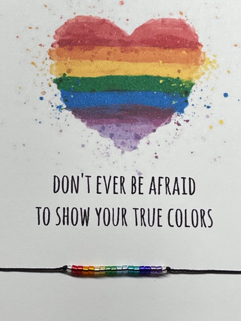 May include: A rainbow heart with a splatter effect and the text "Don't ever be afraid to show your true colors". A rainbow beaded bracelet is below the text.
