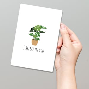 May include: A white card with a green plant in a brown pot and the text "I beleaf in you".