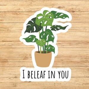 May include: A sticker featuring a potted Monstera plant with large, green, split leaves. The plant is in a tan pot. Below the plant is the text, "I BELEAF IN YOU," in black capital letters.