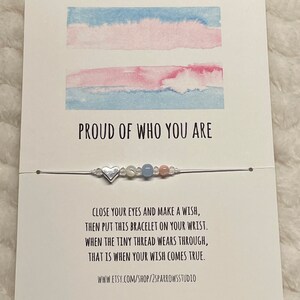 May include: A white card with a watercolor transgender pride flag and the text "PROUD OF WHO YOU ARE". A silver heart charm bracelet with blue, pink, and white beads is on the card. The card also includes the text "CLOSE YOUR EYES AND MAKE A WISH, THEN PUT THIS BRACELET ON YOUR WRIST. WHEN THE TINY THREAD WEARS THROUGH, THAT IS WHEN YOUR WISH COMES TRUE. WWW.ETSY.COM/SHOP/2SPARROWSSTUDIO"