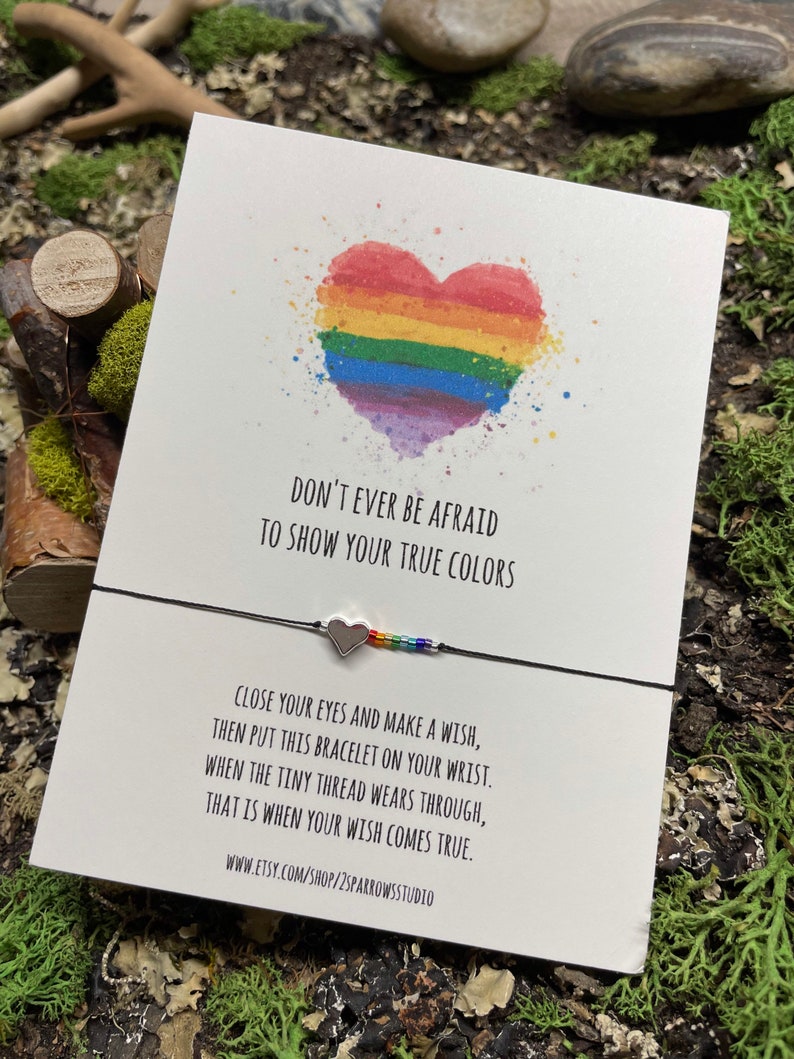 May include: A rainbow heart with a black string bracelet with a silver heart charm. The card reads "Don't ever be afraid to show your true colors. Close your eyes and make a wish, then put this bracelet on your wrist. When the tiny thread wears through, that is when your wish comes true. www.etsy.com/shop/2sparrowsstudio"