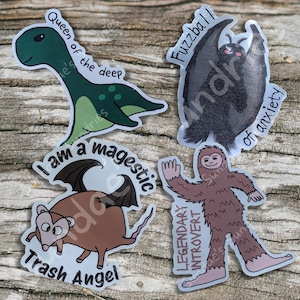 May include: Four whimsical stickers on a wooden surface. The stickers feature cartoon illustrations of a green dinosaur with "Queen of the deep", a black bat with "Fuzzball of anxiety", a brown rat with "Trash Angel", and a brown Bigfoot with "Legendary Introvert".