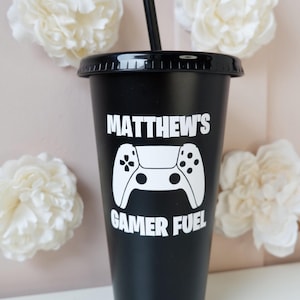 May include: A matte black tumbler with a lid and a black straw. The front features white text that reads MATTHEW'S GAMER FUEL above and below a white graphic of a video game controller. The design is printed directly onto the cup surface.