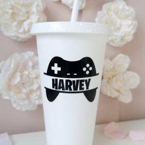 May include: A white plastic tumbler with a matching lid and straw. The front features a black graphic of a video game controller silhouette with the name HARVEY printed in bold black capital letters across the centre of the controller design.