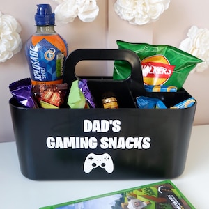 May include: A black plastic caddy with a handle, featuring the white text DAD'S GAMING SNACKS and a game controller icon. The caddy is filled with various snacks and an orange sports drink, with a Minecraft video game case positioned in the foreground.
