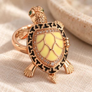 Op de afbeelding: Een roségoudkleurige ring met een schildpadontwerp. Het schildpadschild heeft een lichtgeel midden, zwarte emaille accenten en een rand van kleine heldere steentjes. De kop heeft groene stenen ogen en textuurdetails op het metaaloppervlak.