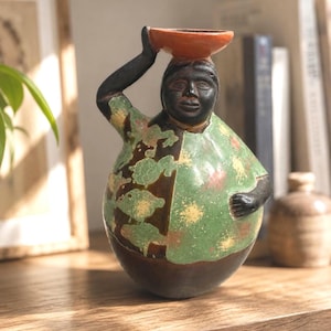 Peut inclure: Une figurine en céramique représentant une personne à la peau foncée, vêtue d'une chemise verte à motifs tachetés clairs. Le personnage a un bras levé et équilibre un petit bol rond de couleur orange sur sa tête. La base est brun foncé.