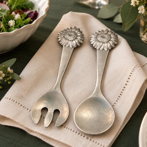 Può includere: Una coppia di posate da insalata in metallo color argento con manici a forma di girasole. Il set include una forchetta da portata a tre rebbi e un cucchiaio, entrambi con finitura opaca e dettagliati disegni di petali sui manici.