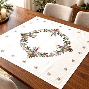 Puede incluir: Un mantel cuadrado blanco con un diseño central de corona bordada compuesto por ramas de pino verdes, velas rojas, campanas doradas y cintas azules. El borde está decorado con pequeños copos de nieve dorados bordados uniformemente.
