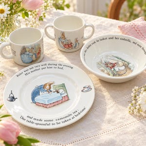 Peut inclure: Un ensemble de vaisselle pour enfants en céramique blanche orné d'illustrations de Pierre Lapin et de texte. L'ensemble comprend deux petites tasses avec anses, un bol peu profond et une assiette. Chaque pièce présente des scènes colorées.