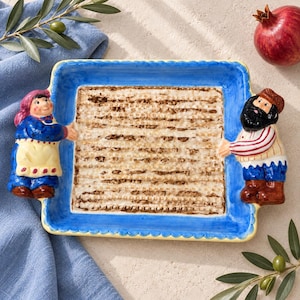 May include: A rectangular ceramic serving tray featuring a central matzah pattern with a blue border. Two sculpted figures, one on each side, hold the tray. The left figure wears a blue dress and apron, while the right figure has a beard and cap.