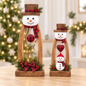May include: Two wooden snowman decorations with arched frames, wearing top hats with red and black buffalo plaid bands. The taller snowman features a hanging plaid ornament, while the shorter one displays a heart ornament and a small snowman figure.