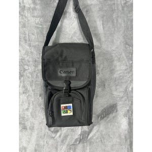 May include: A black Canon camera bag with an adjustable shoulder strap. The bag features a top flap with a plastic buckle closure and a front zippered pocket. A square patch on the front pocket displays the text DV PHOTO PLUS with four icons.