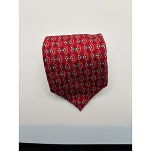 May include: A red necktie featuring a repeating pattern of white dotted arches, small yellow circles, and light blue diamond shapes. The fabric has a smooth texture and the tie is folded into a point at the bottom, displaying a consistent geometric design.