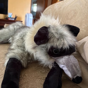 May include: A plush stuffed animal resembling a lemur with long, shaggy cream and grey fur, black ears, black eye patches, and black limbs. The creature has a pointed grey snout with a small black nose and dark, round, glossy eyes.