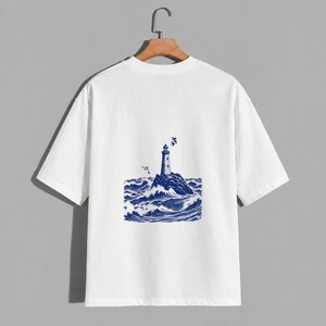 Può includere: Una t-shirt bianca a maniche corte con una stampa grafica blu al centro del petto. L'illustrazione raffigura un faro su uno sperone roccioso circondato da onde oceaniche agitate, con un delfino e uccelli nel cielo sovrastante.