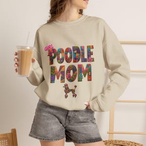 May include: A beige crewneck sweatshirt featuring the text GIRL POODLE MOM in large, multi-colored, floral-patterned block letters. Below the text is a small, matching floral-patterned poodle graphic. The sweatshirt has long sleeves and a ribbed collar.