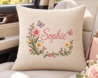 May include: A square beige throw pillow featuring an embroidered floral wreath with pink, yellow, and white flowers, green leaves, and two butterflies. The name Sophie is embroidered in pink cursive script in the centre of the floral arrangement.