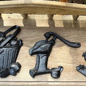 May include: A collection of black cast iron decorative pieces, including a teapot, coal buckets, a water pump, and a stove. The pieces are arranged on a wooden surface, showcasing a rustic, vintage aesthetic.