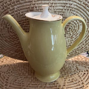 May include: A yellow ceramic teapot with a white lid and gold trim. The teapot has a rounded body and a spout on one side. The handle is on the opposite side of the spout.