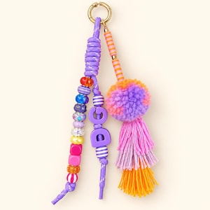 May include: A keychain featuring a gold-tone metal ring, two purple cord strands, and a multi-colored tassel. One cord holds various textured beads and the letters D and G in purple. The other cord has orange and pink striped beads and a pom-pom.