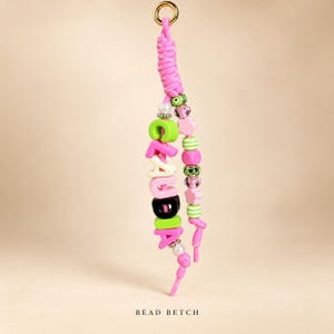 Può includere: Un portachiavi decorativo con un anello in metallo dorato attaccato a un cordino intrecciato rosa. Un filo mostra il nome CLAUDIA in lettere colorate, mentre l'altro filo presenta una varietà di perline rosa, verdi e bianche.