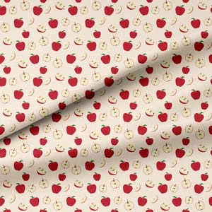 May include: A fabric featuring a repeating pattern of red apples and light beige apple slices on a cream-colored background. The design includes whole red apples with green stems and cross-sectioned apple slices in a light beige tone.