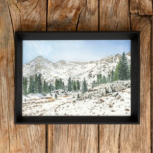 May include: A framed watercolor painting of a snowy mountain landscape with a black frame. The painting depicts a mountain range with snow-capped peaks and a forest of pine trees in the foreground.