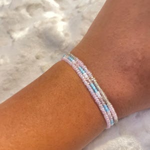 May include: A multi-strand bracelet composed of three rows of small seed beads. The beads feature a color palette of light pink, sky blue, and iridescent white, arranged in a delicate, thin design that wraps around the wrist.