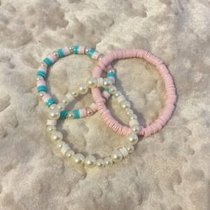 May include: Three stacked bracelets featuring a mix of materials. One bracelet consists of light pink clay heishi beads. Another features turquoise, white, and pink clay discs with small white pearls. The third is made of white pearls and white discs.
