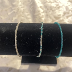May include: Three thin beaded bracelets arranged in a row. The first bracelet is made of clear, translucent beads. The second bracelet features a mix of clear and turquoise beads. The third bracelet consists entirely of solid turquoise beads.