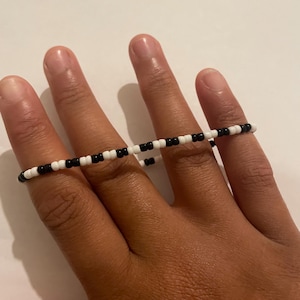 May include: A thin, circular ring constructed from small, alternating black and white seed beads. The beads are strung together in a repeating pattern, creating a simple, lightweight band that encircles the finger with a uniform, monochromatic design.