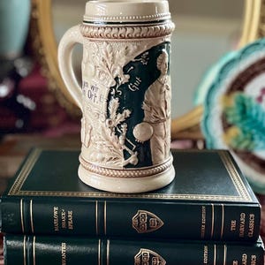 May include: A beige and green ceramic beer stein with a handle, featuring relief carvings of figures and text. The stein is placed on top of three dark green hardcover books with gold lettering. The text "Gut" is visible on the stein.