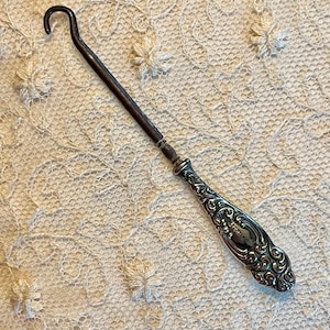 May include: An antique silver-toned button hook with a curved hook end and an ornate handle. The handle features a detailed floral design. The button hook is approximately 7 inches long.