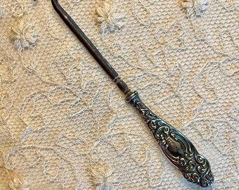 May include: An antique silver-toned button hook with a curved hook end and an ornate handle. The handle features a detailed floral design. The button hook is approximately 7 inches long.
