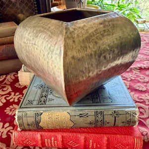 May include: A large, hammered metal bowl with a heart-like shape, resting on a stack of antique books. The bowl has a textured, metallic finish. The books are bound in red and blue, with gold lettering.