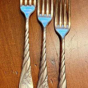 May include: Three antique silver forks with twisted handles and ornate floral designs. The tines are gold-toned, and the handles have a detailed, rope-like twist. The forks are arranged on a wooden surface, showcasing their vintage charm.